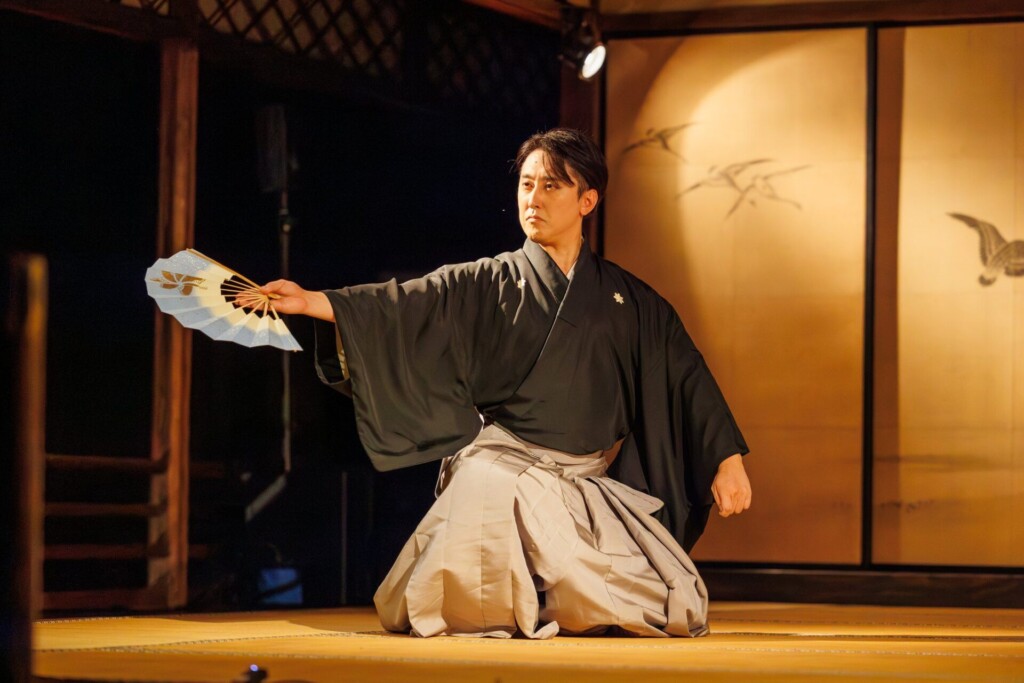 扇を手に着物姿で舞台に座る男性の踊り手